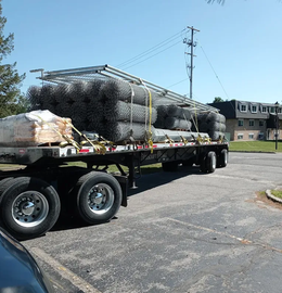 Flatbed trailer loaded with bundled black wire mesh rolls in a parking lot under a blue sky