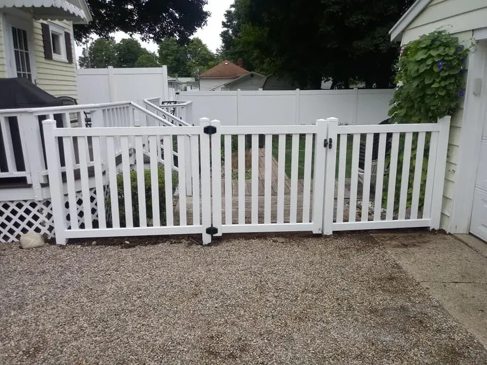 White backyard fence gate with gravel driveway and greenery beside a house