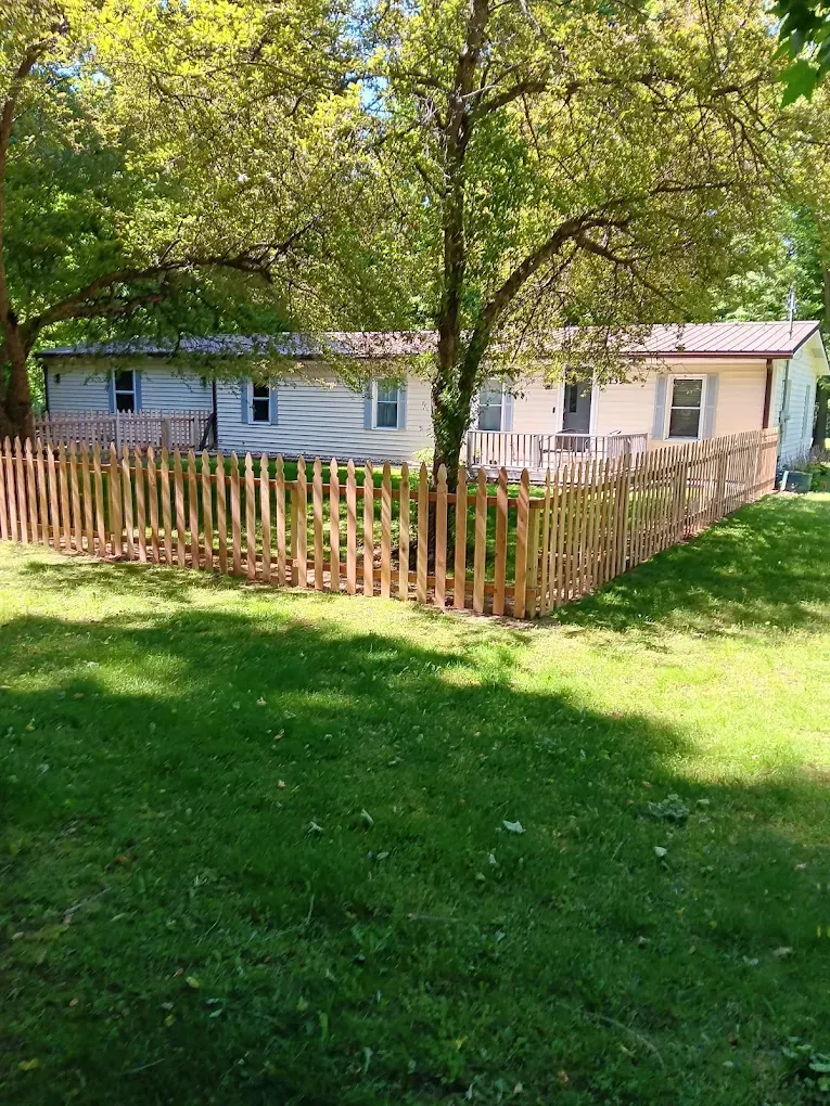 White house behind a wooden picket fence, shaded by large trees, with a grassy yard in front