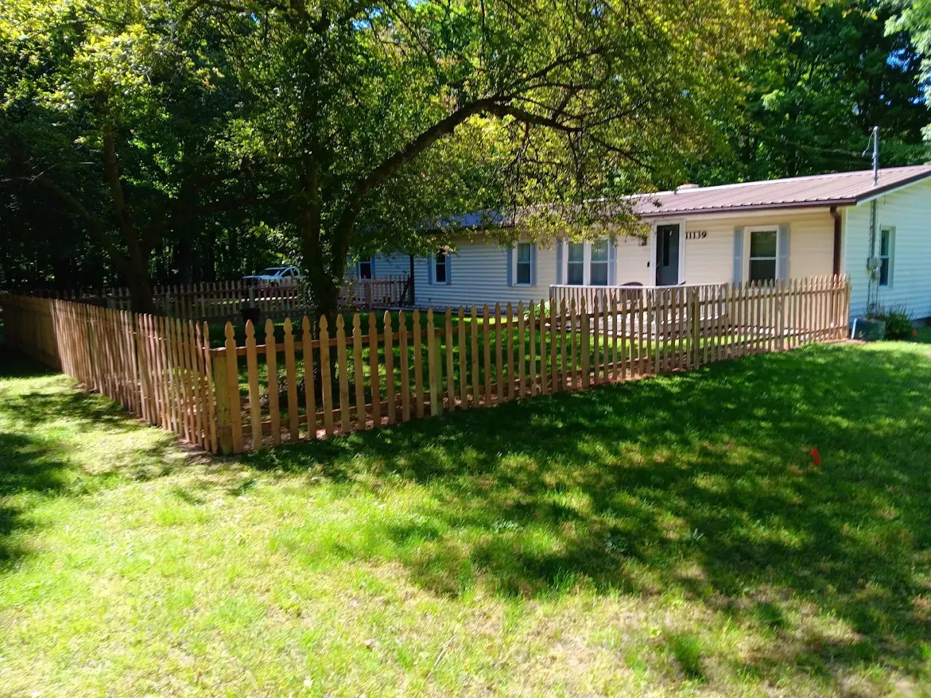 Small white house with a wooden fence, shaded by trees, beside a sunny green yard.