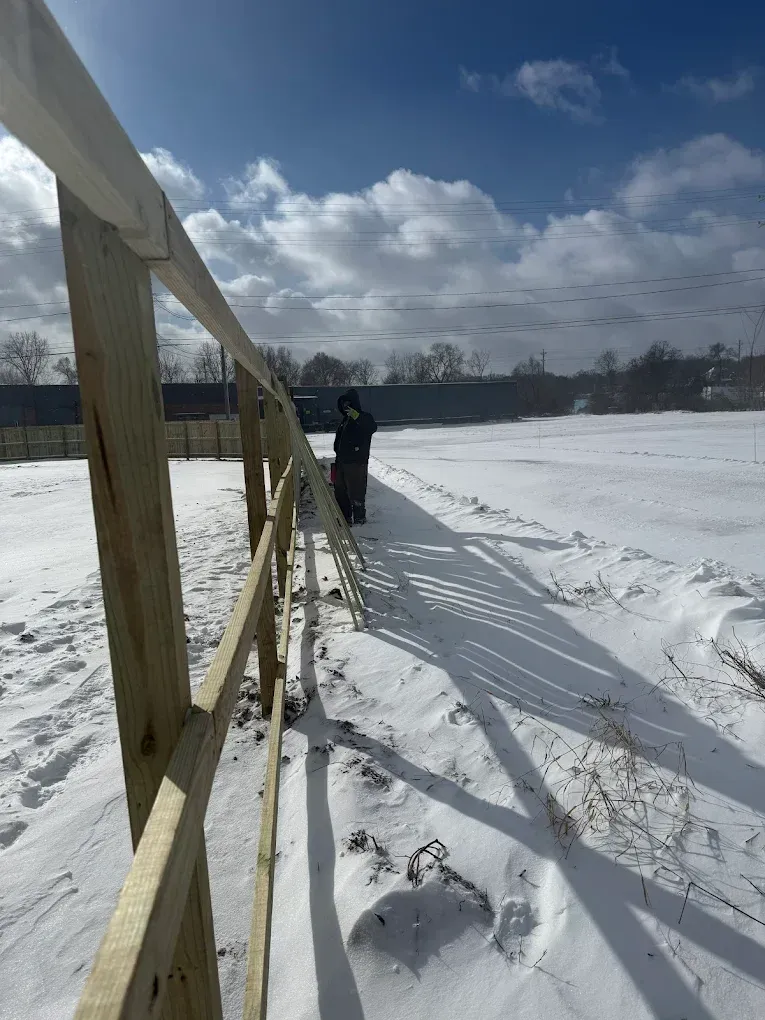 Person walking on a snowy path beside a yellow fence under a cloudy sky