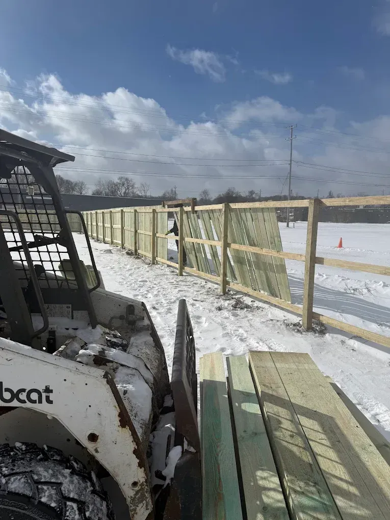 Snowy construction site with a skid steer beside a fenced walkway under a cloudy sky
