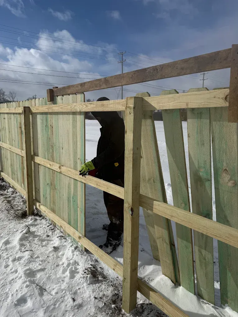 Snow-covered wooden fence with a broken opening and a dark dog standing in the gap