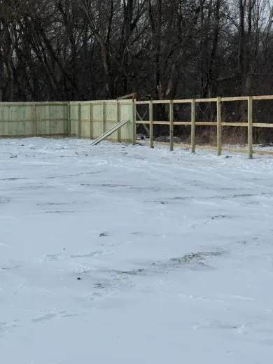 Snow-covered yard with a wooden fence and leafless trees in the background