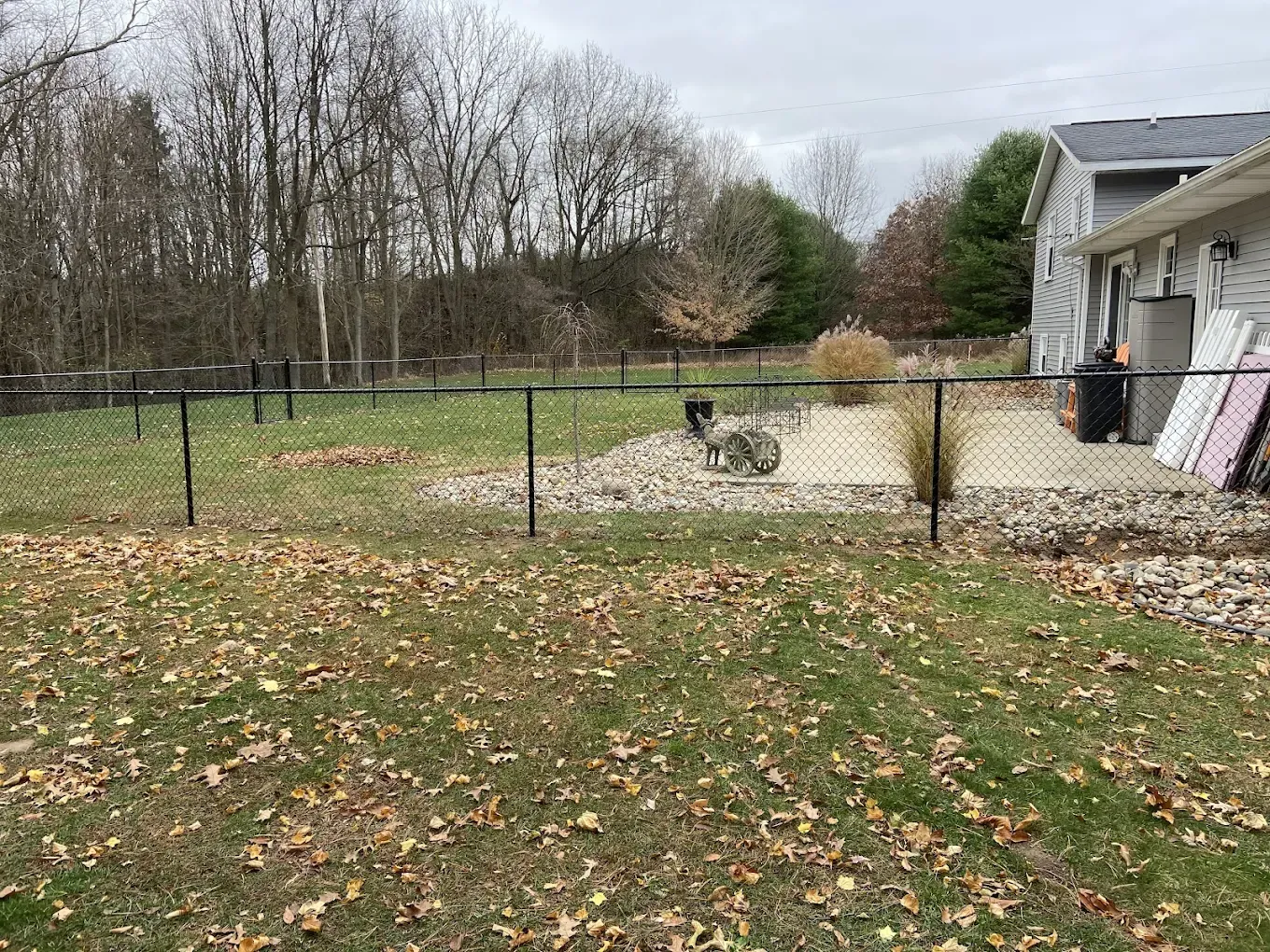 Backyard with leaf-covered grass, fenced pond, and a house at right under cloudy skies