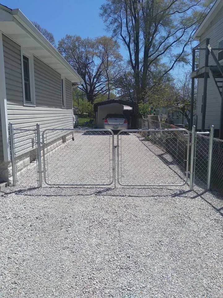 Gravel driveway between two houses with chain-link gate, leading to a carport at the back
