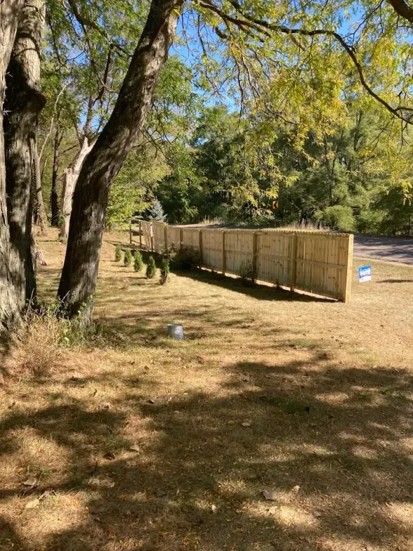 Wooded park path beside a long wooden fence under leafy trees, with dry grass and dappled sunlight.
