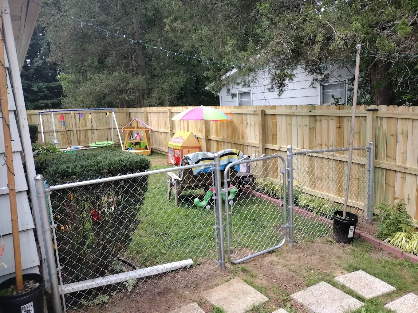 Backyard with chain-link gate, wooden fence, hedges, patio pavers, and outdoor furniture under trees