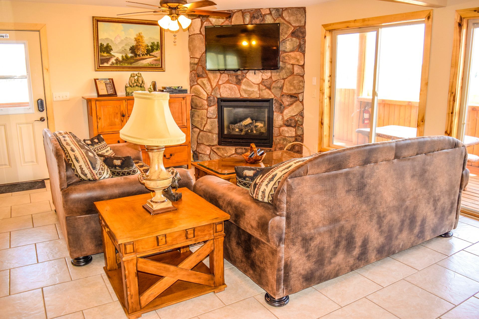 Living room with brown leather sofas, stone fireplace with TV, wooden side table, and sliding glass door.