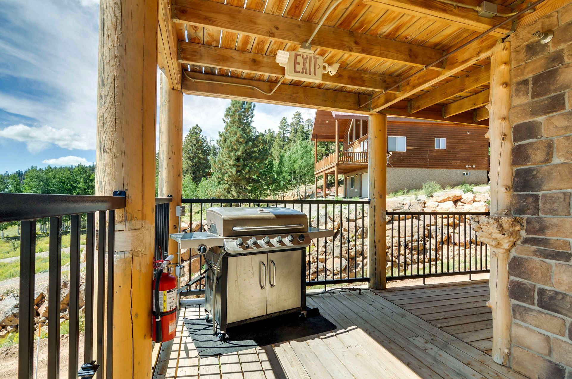 Outdoor wooden deck with a grill and a fire extinguisher. The view includes trees, another building, and a blue sky.