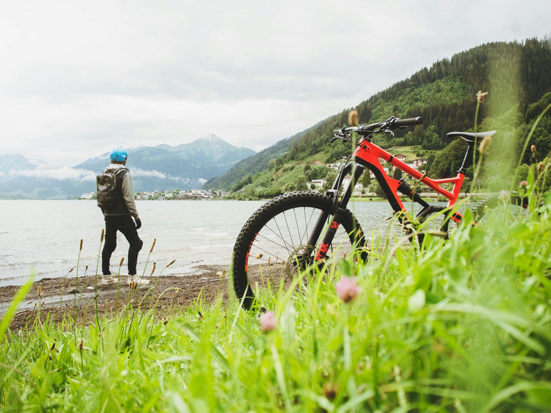 Person with a backpack stands at the edge of a lake, mountain bike in the foreground, mountain range in the background.