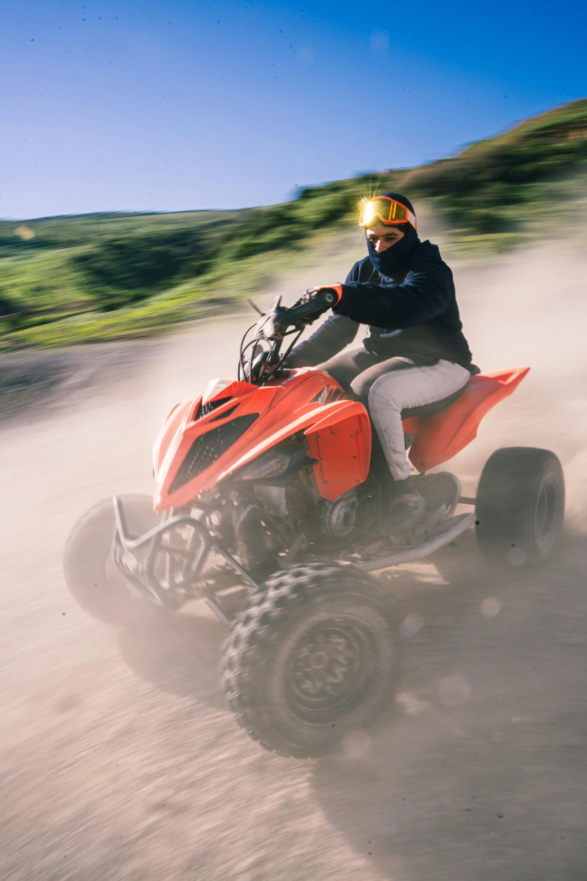Person riding a red ATV on a dirt path, kicking up dust. Green hills and blue sky in the background.