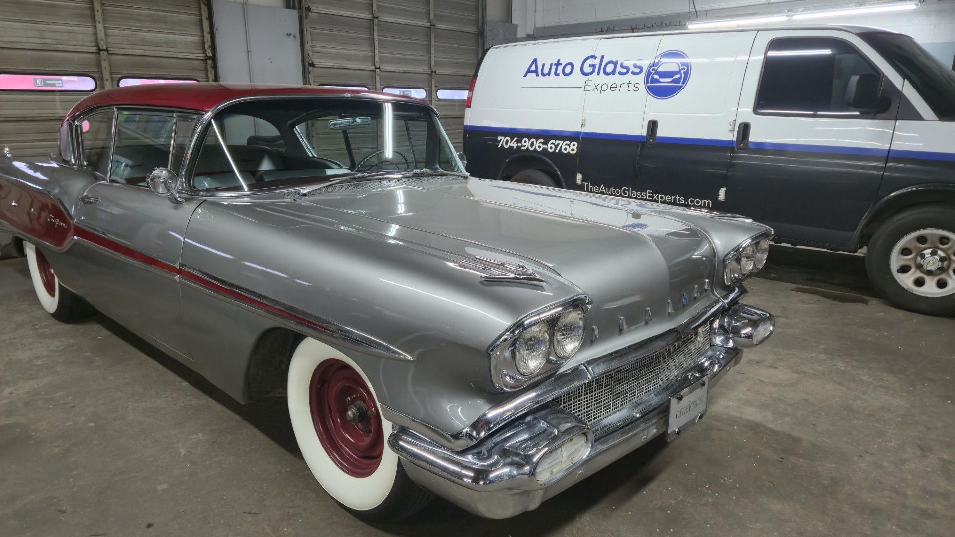 Classic silver and red vintage car parked indoors beside a blue and white van.