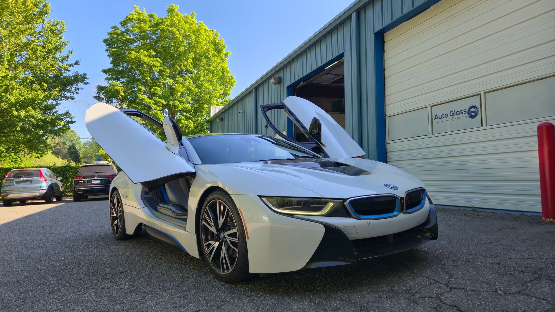 White sports car with scissor doors open, parked outside an industrial building on a sunny day