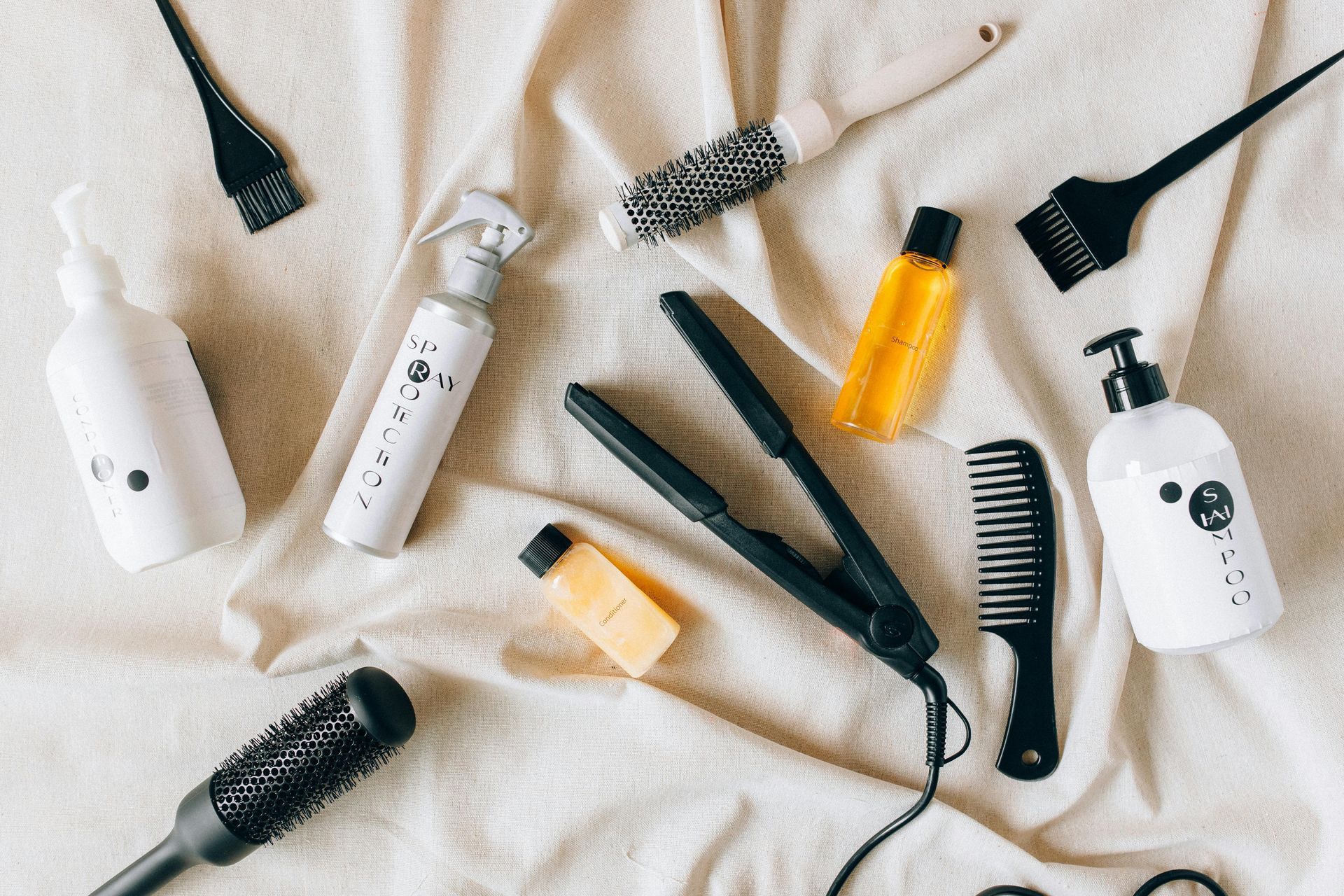 Hair styling tools on a white cloth, including a flat iron, brushes, spray bottle, and bottles of products.