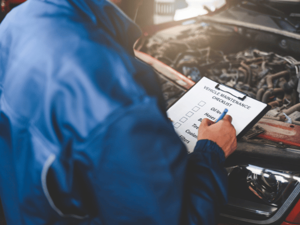 Mechanic in blue jacket writing on a checklist beside an open car hood in a garage