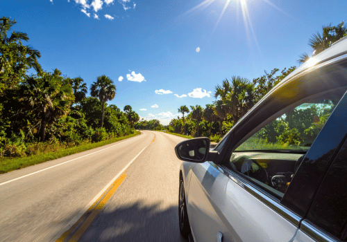 White car driving along a sunny tropical road lined with palm trees under a bright blue sky.