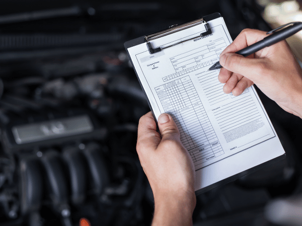 Hands writing on an auto repair checklist clipboard beside a car engine