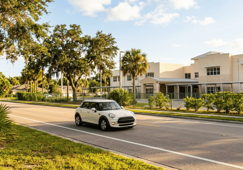 White MINI Cooper driving past a school on a sunny day, highlighting back-to-school