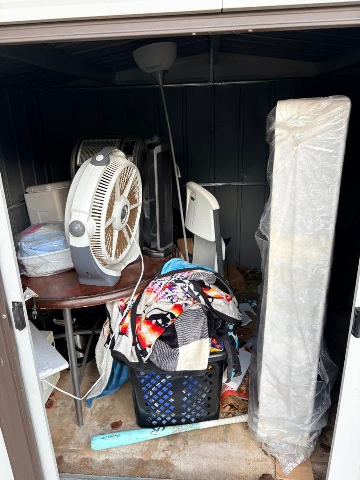 Cluttered storage nook with a fan, baskets, boxes, and a tall wrapped item leaning against the wall