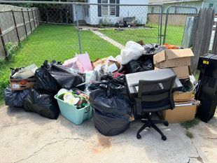 Overflowing trash piled on a driveway beside a green backyard and chain-link fence.