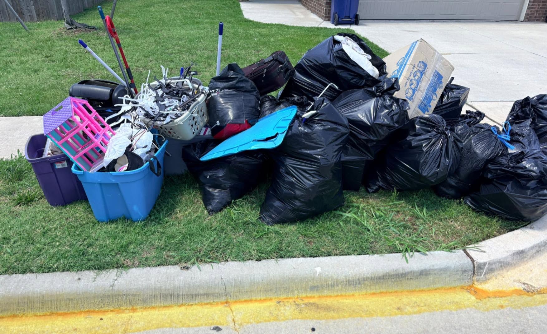 Pile of black trash bags and assorted debris on a curb beside a grassy lawn.