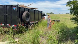 Black utility trailer and white truck parked in a grassy field, with a person working beside them under a blue sky