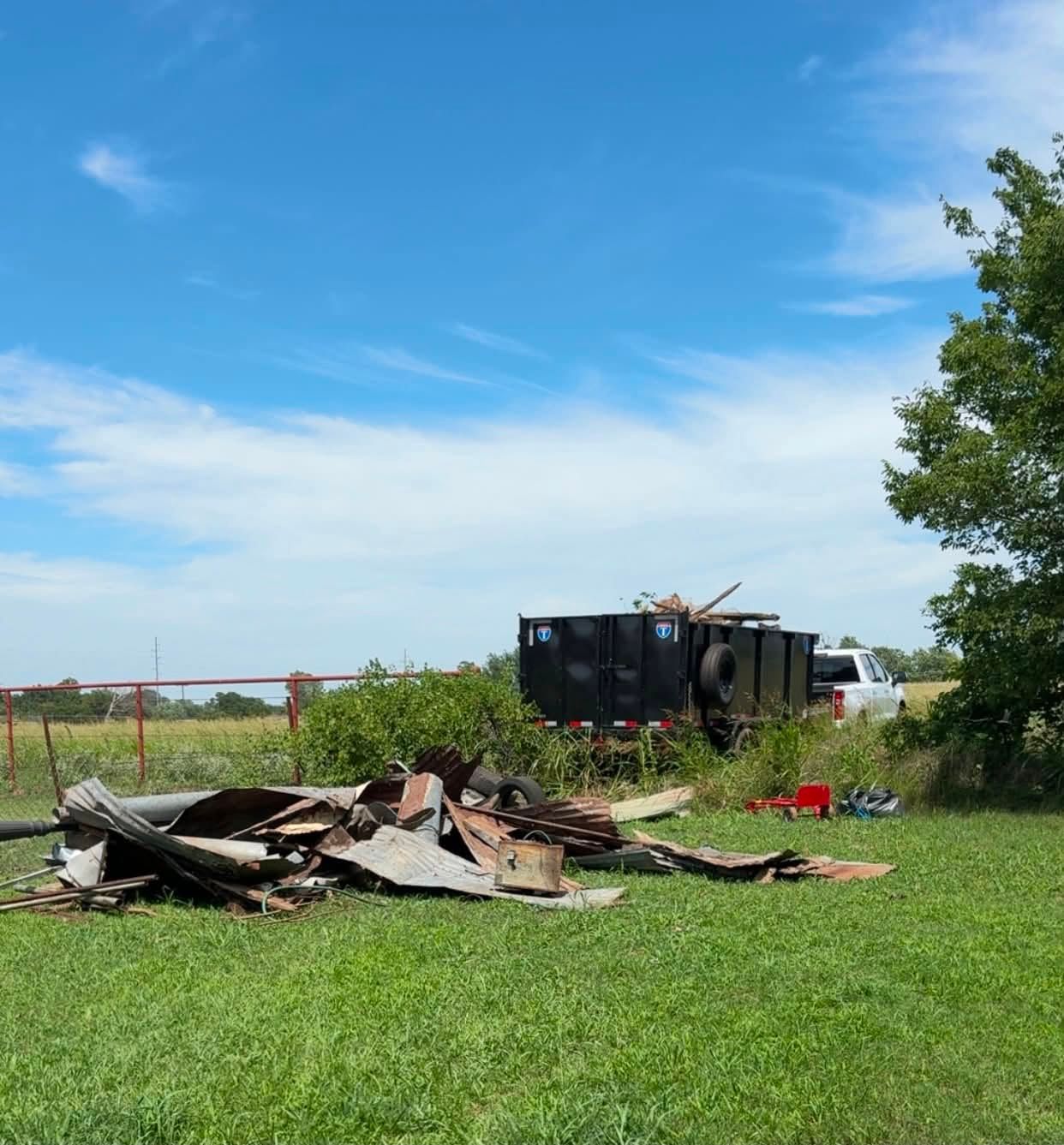 Burned debris pile in a grassy field beside a tipped black trailer under a blue sky