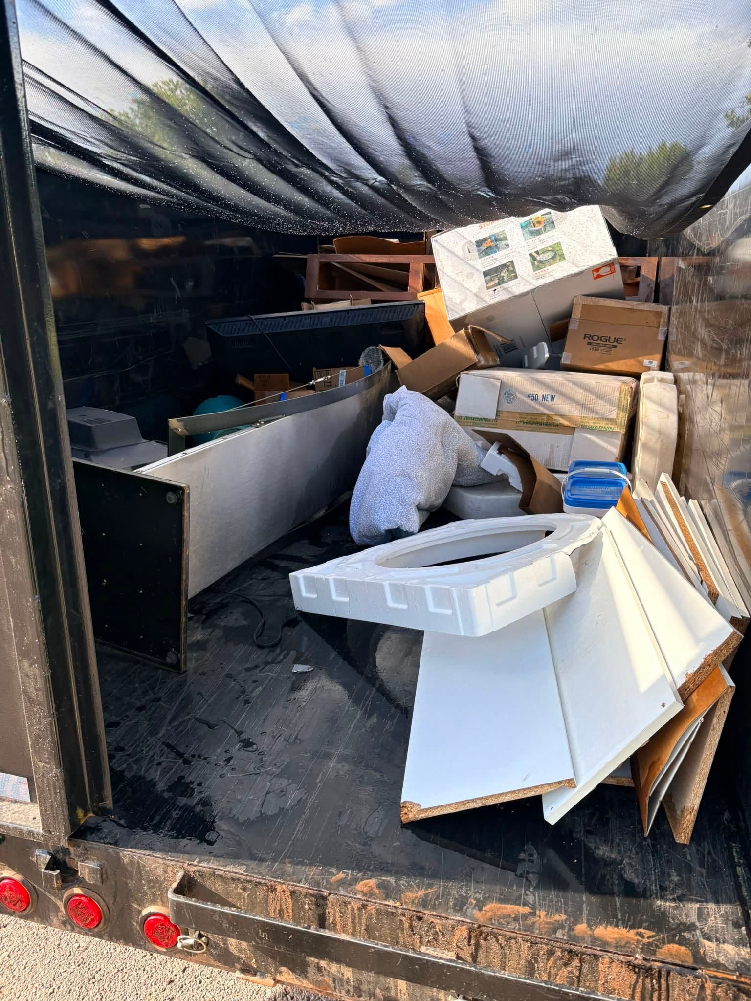 Cluttered moving trailer packed with boxes, furniture, and loose items under a black tarp.