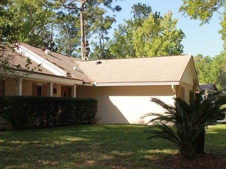 Beige house with a tan roof, green lawn, and trees in the background under a blue sky.