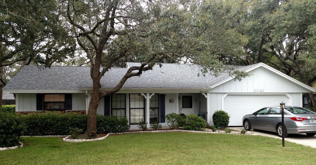 White house with gray roof, green lawn, car parked in driveway, and large tree in the front yard.