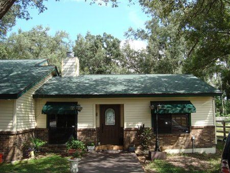 Tan house with green roof, awnings, and trees in background. Brick accents, brown door.