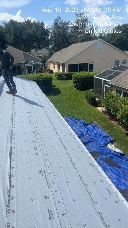 Person on a roof with a white underlayment, houses and greenery visible on a sunny day.