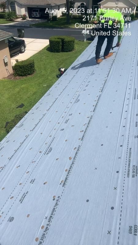 Workers installing roofing material on a house. Blue underlayment, green grass, and residential setting.