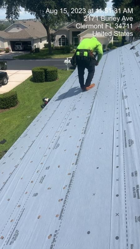 Roofer on a gray roof, wearing a safety vest and harness. Clermont, FL. August 15, 2023.
