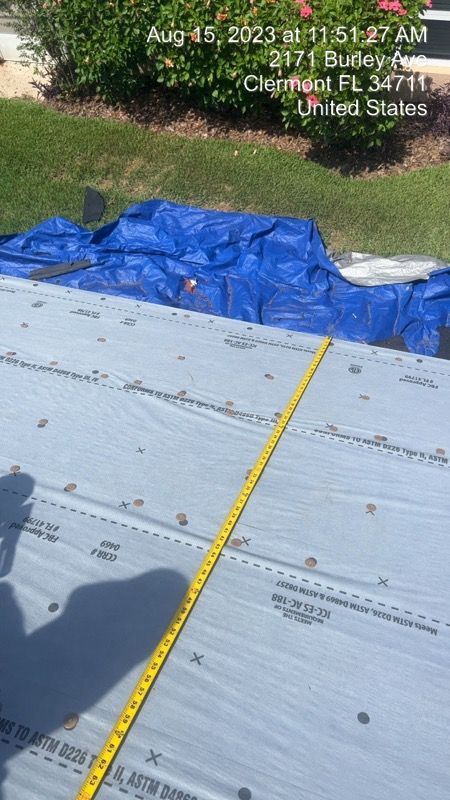 A measuring tape extends across a gray roof material with a blue tarp in the background.