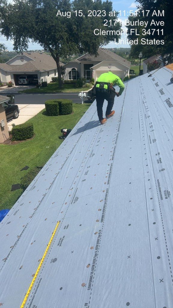 Roofer on a gray metal roof, wearing safety gear. Green lawn and houses in background.