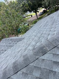 Gray asphalt shingle roof on a house, angled view, with trees and a street visible in the background.