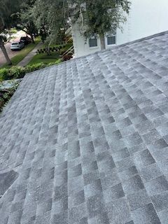 Gray asphalt shingle roof with a house and trees in the background on a sunny day.