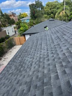 Dark gray asphalt shingle roof on a house, with a cloudy sky and green trees in the background.
