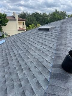 Gray shingle roof with skylight, black bucket. A building is visible in the background. Cloudy sky.