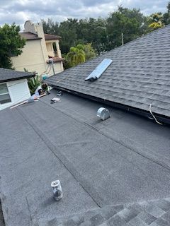 Dark gray asphalt shingle and flat tar roof with vents, under cloudy sky. Buildings and trees in background.