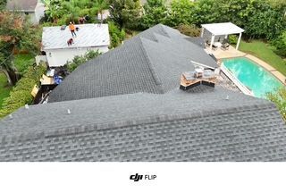 Workers on a roof with a gray shingle installation; house, yard, and swimming pool visible.