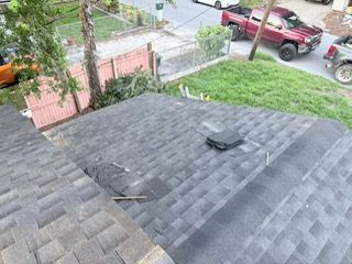 Overhead view of a dark gray shingled roof with a vent, next to a grassy area with vehicles and a fence.