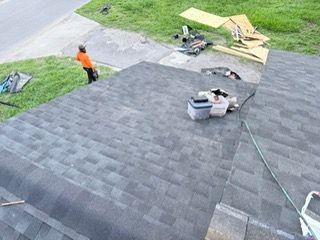 Person in orange shirt on a roof, working on dark gray shingles. Construction debris and green grass visible.
