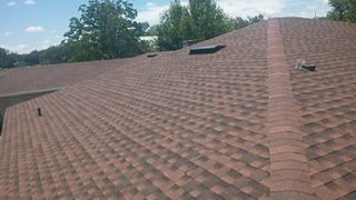 Brown asphalt shingle roof on a house, angled view, with trees in the background under a blue sky.