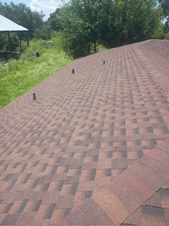 Brown asphalt shingle roof, with dark patches, vents, and green trees in background.