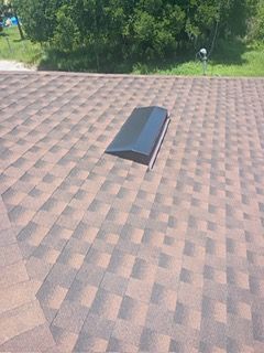 Brown shingled roof with a dark rectangular skylight. Green trees and grass in the background.