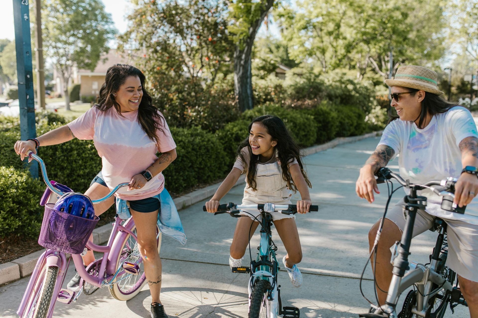 Three people smiling while riding bicycles along a sunny, tree-lined path.