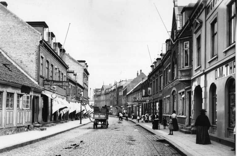 Historisk sort-hvid gadescene med hestevogn på en brostensbelagt vej, omkranset af butikker og huse